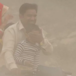 man driving a motorbike through a smog filled street with a child while shielding the child's face from the smog